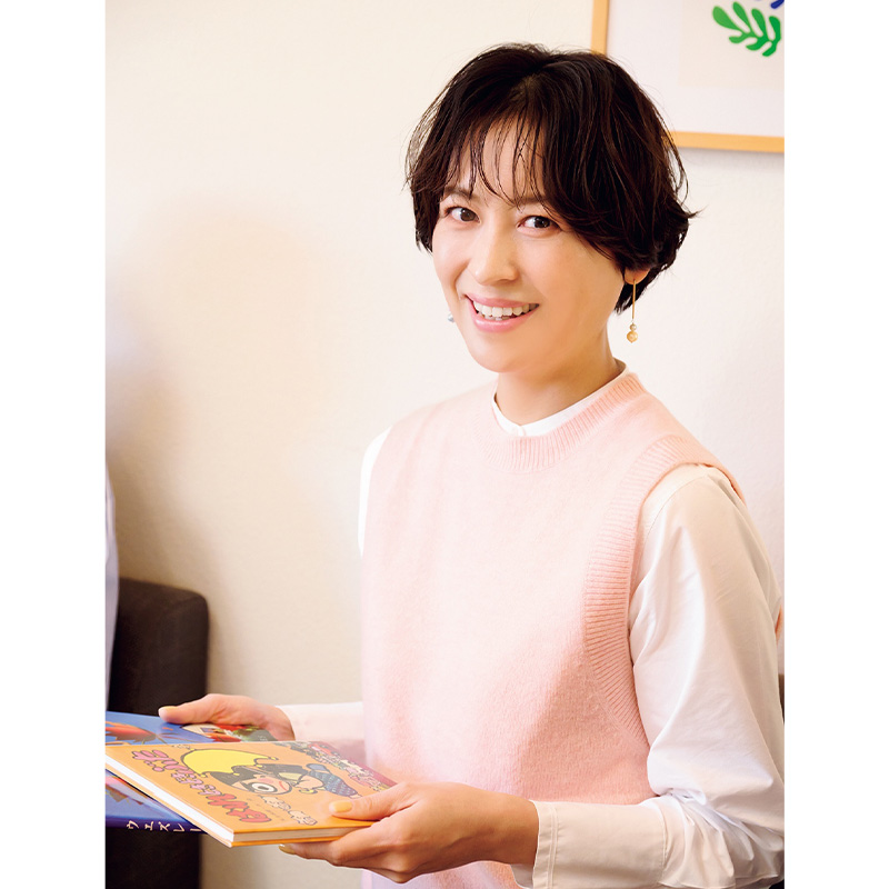 児童書を手に微笑む白Tシャツにピンクのベストを着た青木裕子さん