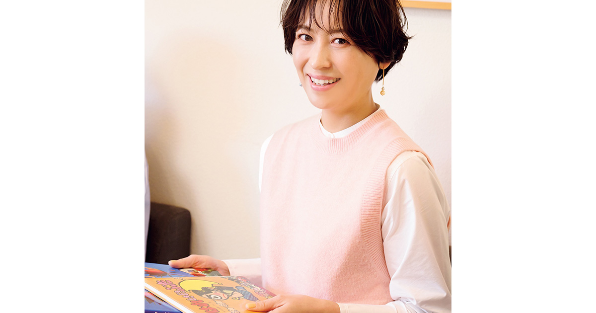 児童書を手に微笑む白Tシャツにピンクのベストを着た青木裕子さん