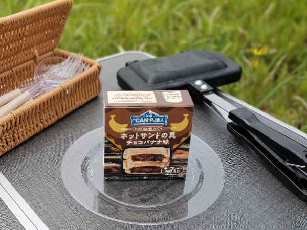 キャンプ用テーブルの上にある「K&K ”CAN”P の達人 ホットサンドの具チョコバナナ味」とホットサンドメーカーが並んでいます。