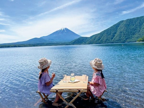 キャンプ中に、木製のテーブルとイスを本栖湖の浅瀬に置き、湖に足までつかりながら座って冷たい飲み物を飲む幼い姉妹